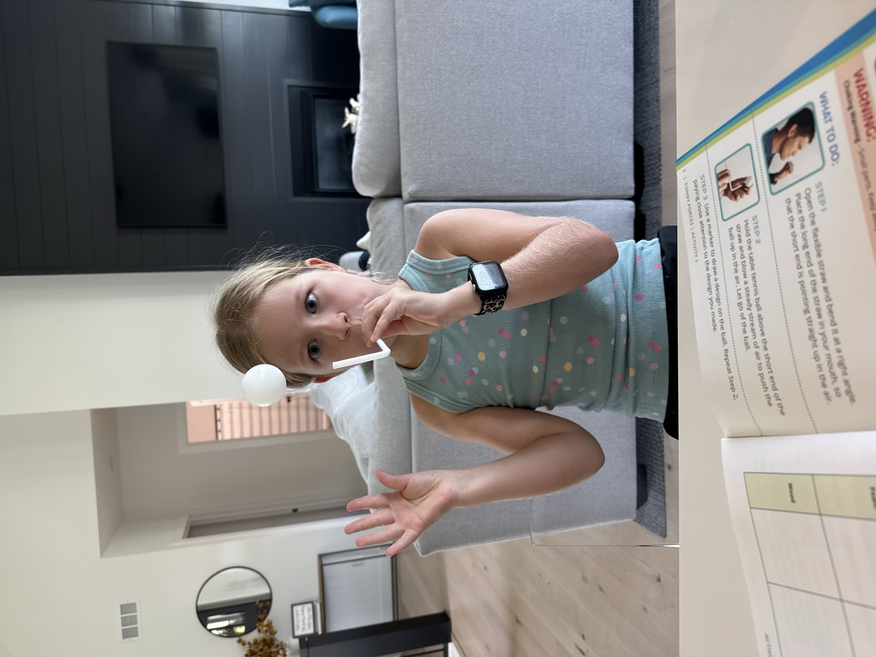 Scarlett blowing through a bent straw to hold a white ball in the air, workbook open on the table